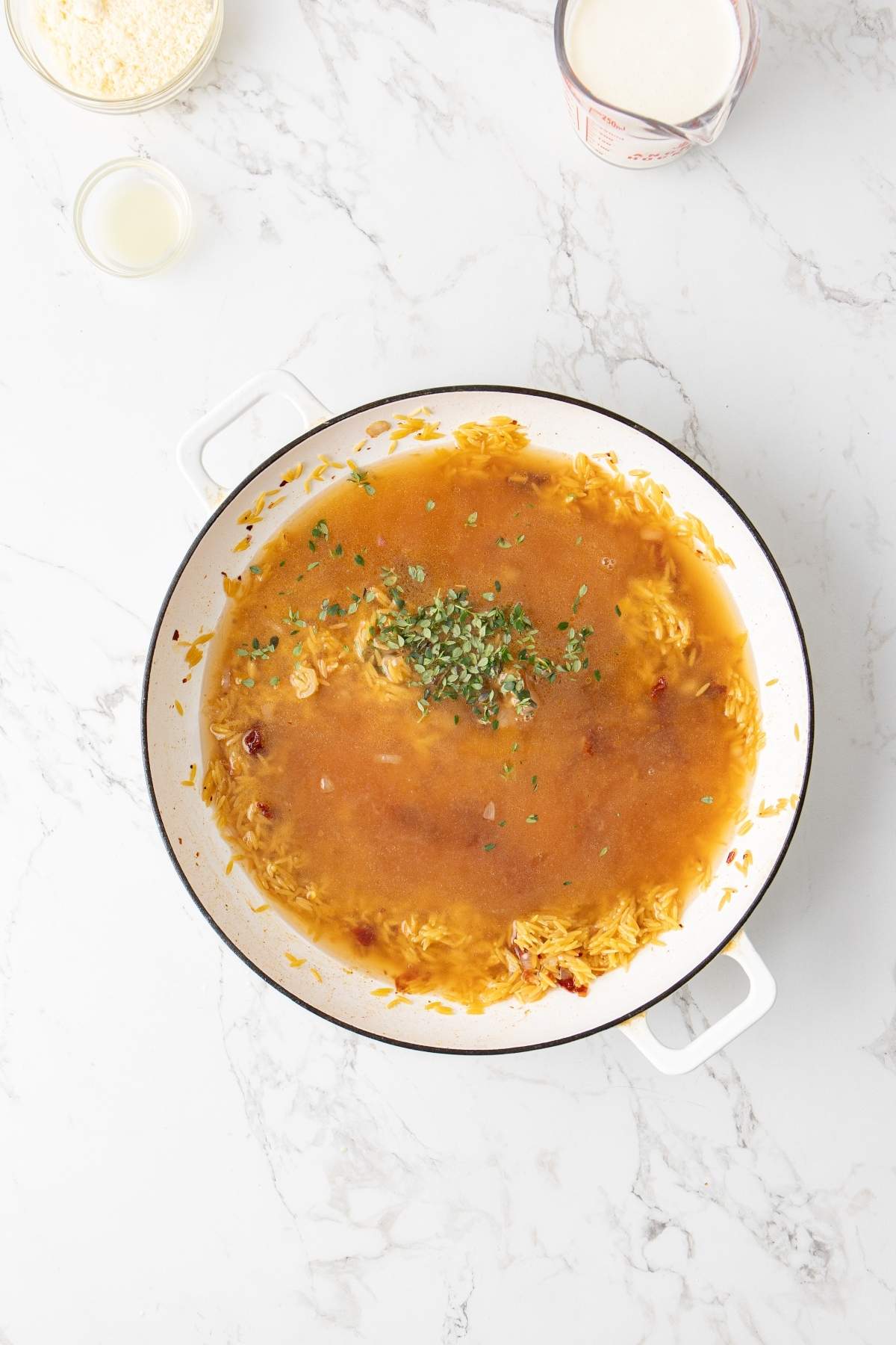 A white skillet with cooked risotto in brown liquid, topped with chopped fresh herbs. Cream and grated cheese are in containers next to the skillet on a white marble surface. For an Italian twist, swap risotto for flavorful chicken orzo.