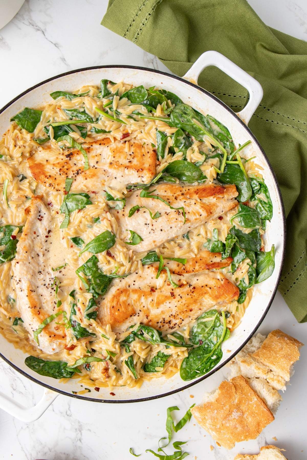 A skillet dish featuring Italian chicken orzo with seared chicken breasts, creamy orzo pasta, and fresh spinach. A green napkin and pieces of bread are on the side.